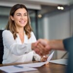 An insurance agent sitting at her desk with paperwork on it while shaking hands with a client