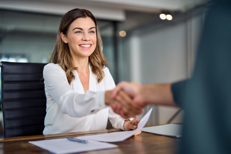 An insurance agent sitting at her desk with paperwork on it while shaking hands with a client